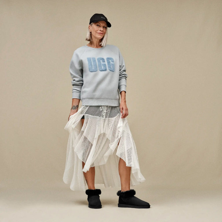 woman wearing a gray UGG sweater with 'UGG' branding, a white skirt, and black shoes on a beige background.