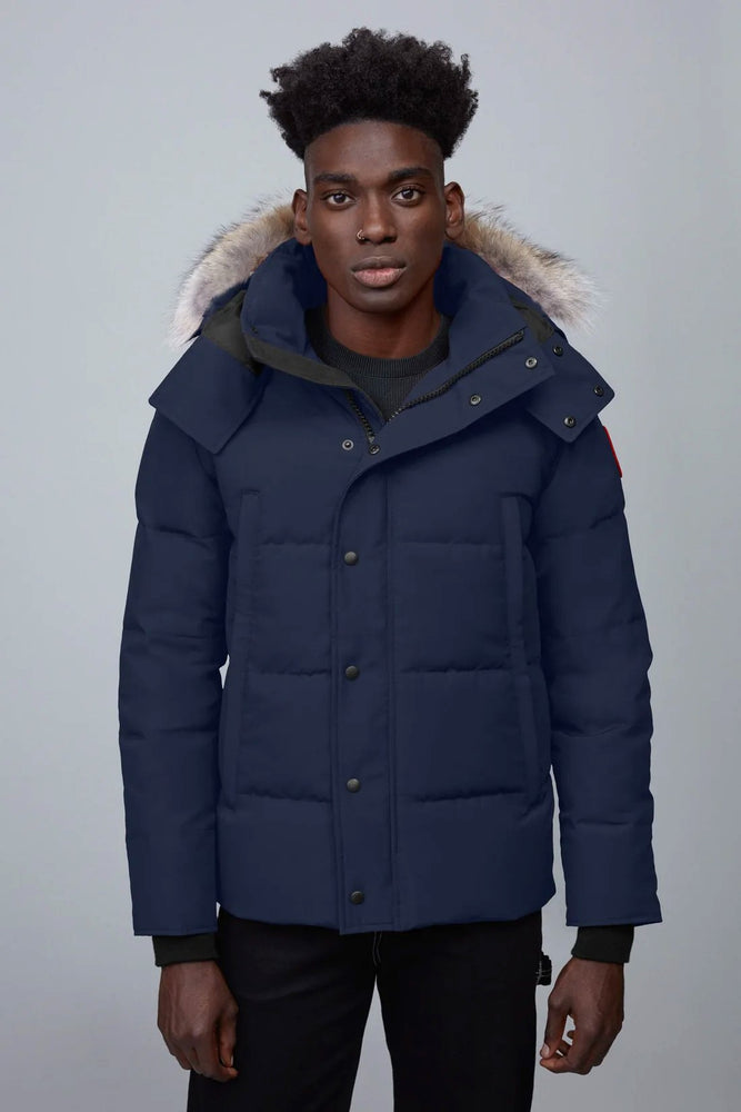 Man wearing a blue winter Canada Goose Wyndham Parka Goose Men's Winter Jacket at Volonka. coat with fur-lined hood against a gray background