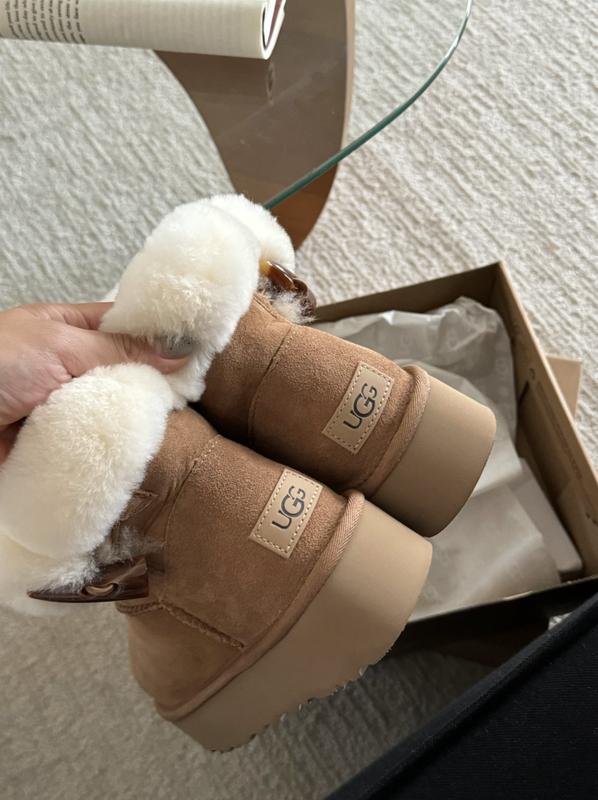Brown UGG boots with white fur lining held above an open box on a light-colored surface.