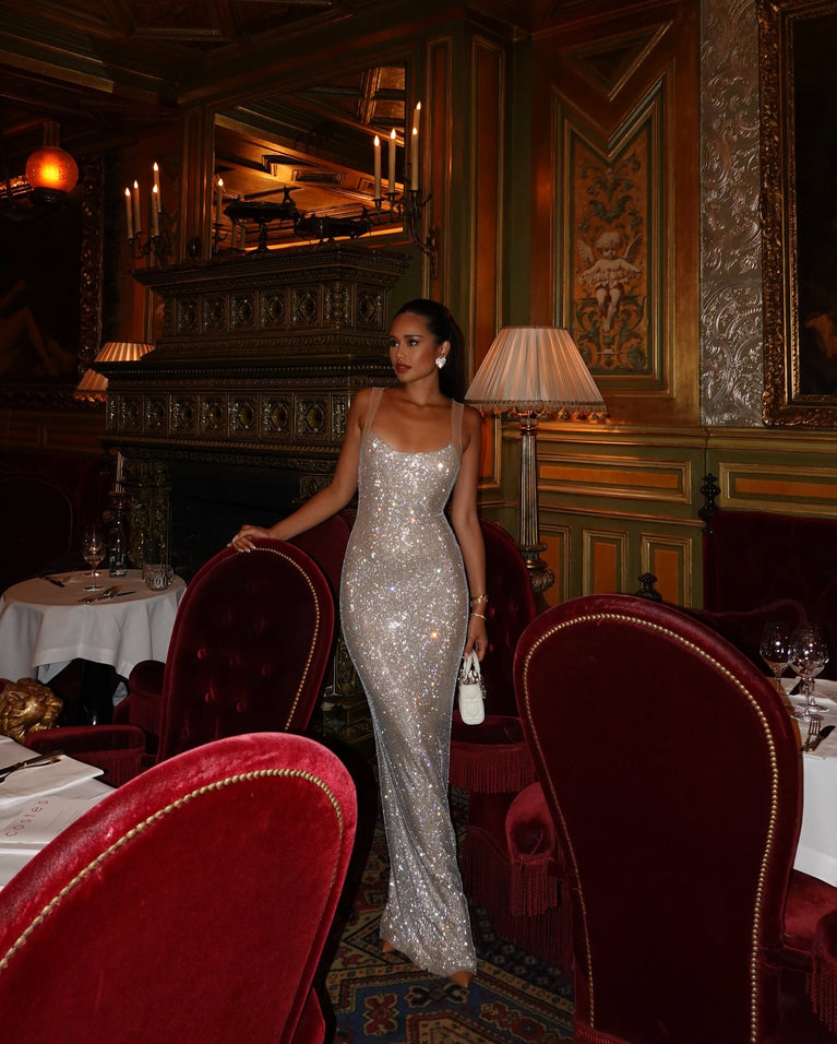 Elegant woman in a backless sparkly maxi evening dress standing in a lavish restaurant setting.