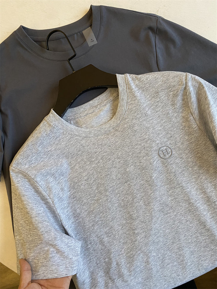 Gray t-shirt with a logo held by a hand, with a black t-shirt in the background.