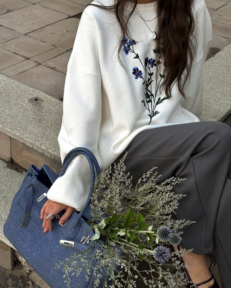 Person wearing a white sweater with floral embroidery, holding a blue handbag and a bouquet of flowers.