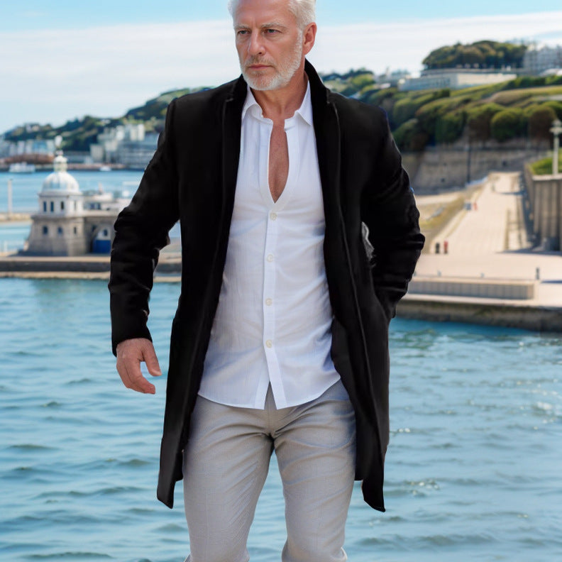 Man in a black coat and white shirt standing by a waterfront with greenery and buildings in the background.