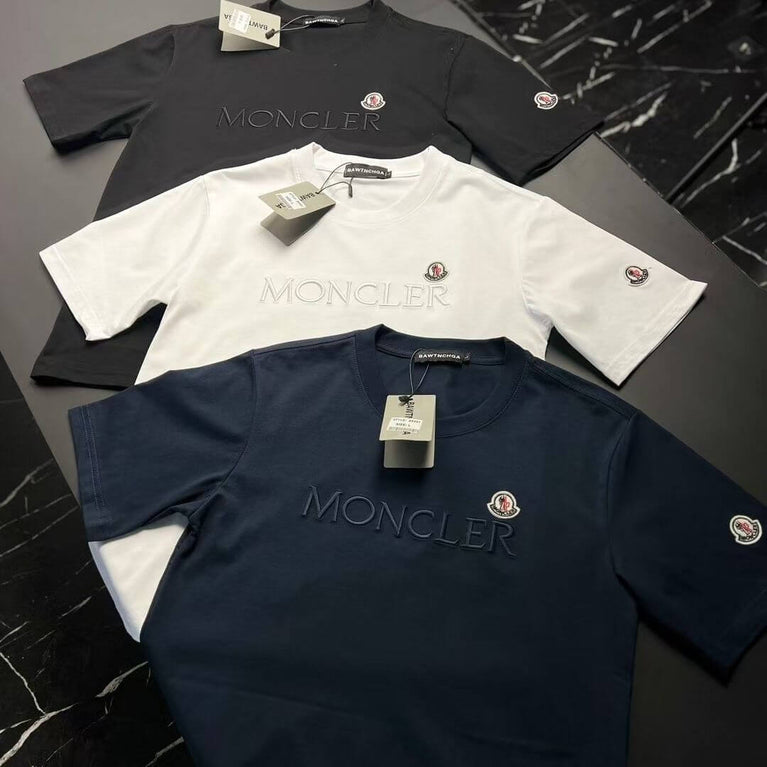 Three Moncler t-shirts in black, white, and navy on a dark surface.