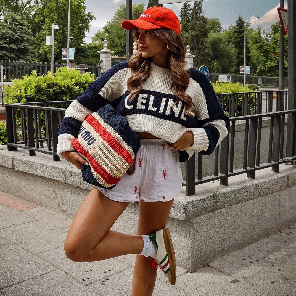 Woman wearing a Celine sweater and red cap, holding a striped bag on a city street.