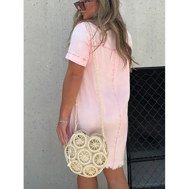 Woman wearing a pink denim dress holding a round woven bag against a gray wall.