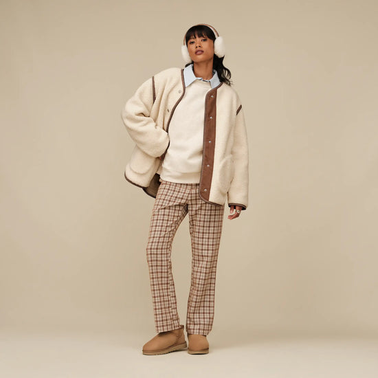 woman wearing a cream jacket with brown accents, plaid pants, and beige boots on a beige background.