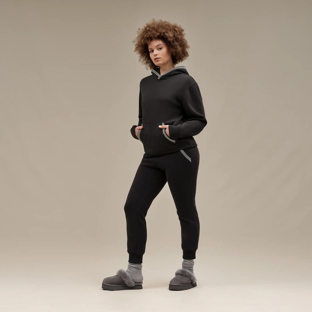 Person wearing a black hoodie and pants with gray slippers on a beige background