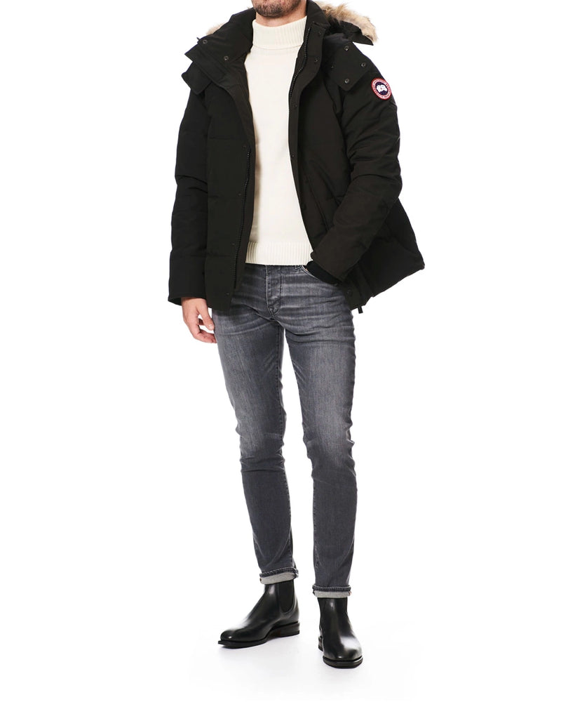 man wearing a black coat with fur hood, white shirt, gray jeans, and black boots on a white background. Canada Goose Wyndham Parka with Fur jacket