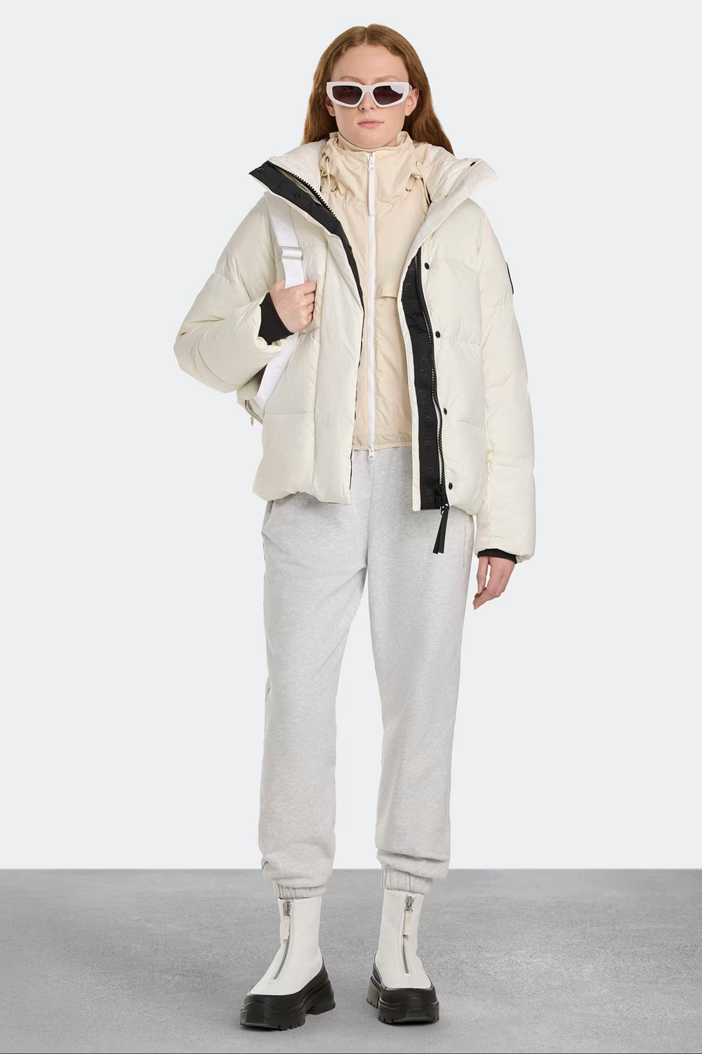 Woman wearing a cream jacket with black accents, light gray pants, and black shoes. Canada Goose Junction Parka Black Label Unisex Winter Jacket at Volonka store.