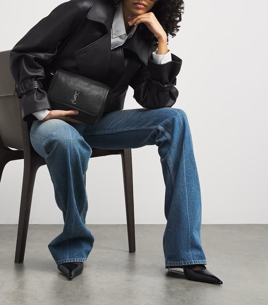 Person sitting on a chair wearing a black leather jacket, blue jeans, and holding a black handbag. Saint Laurent Niki Small leather messenger bag at Volonka.