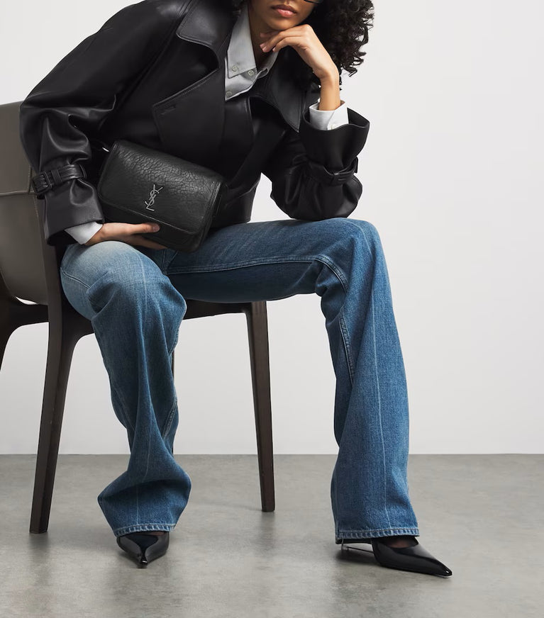 Person sitting on a chair wearing a black leather jacket, blue jeans, and holding a black handbag. Saint Laurent Niki Small leather messenger bag at Volonka.
