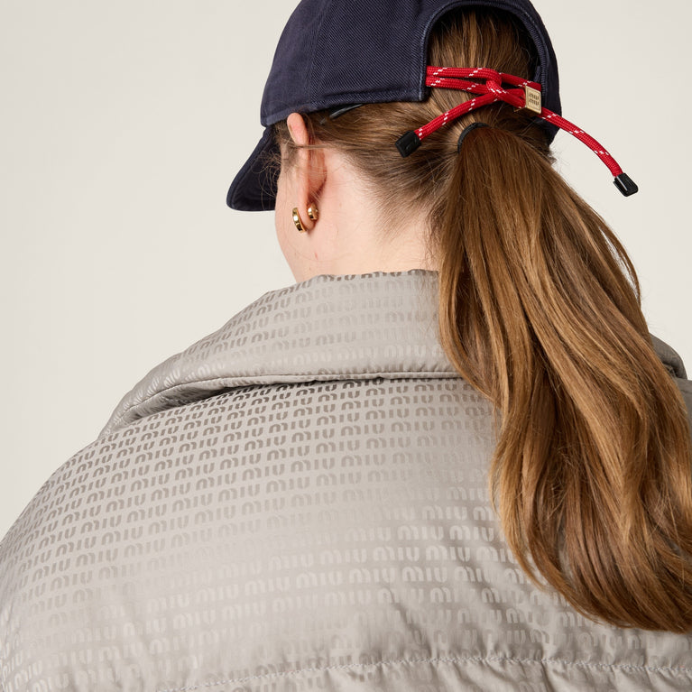 Person wearing MIUMIU a navy cap with a red hair tie, beige jacket, and gold earring on a neutral background