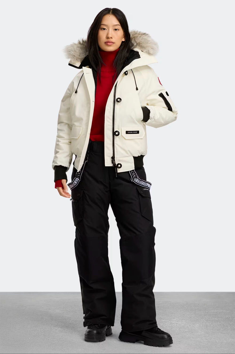 woman wearing a white winter jacket with fur hood and black pants on a light gray background