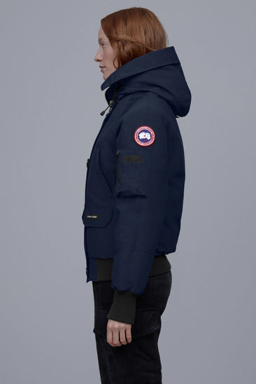 Person wearing a navy blue coat with a brand logo on a gray background. Canada Goose Chilliwack Bomber Women's Winter Hooded Jacket
