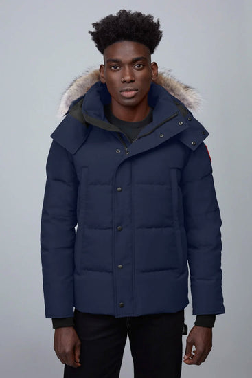 Man wearing a blue winter Canada Goose Wyndham Parka Goose Men's Winter Jacket at Volonka.  coat with fur-lined hood against a gray background