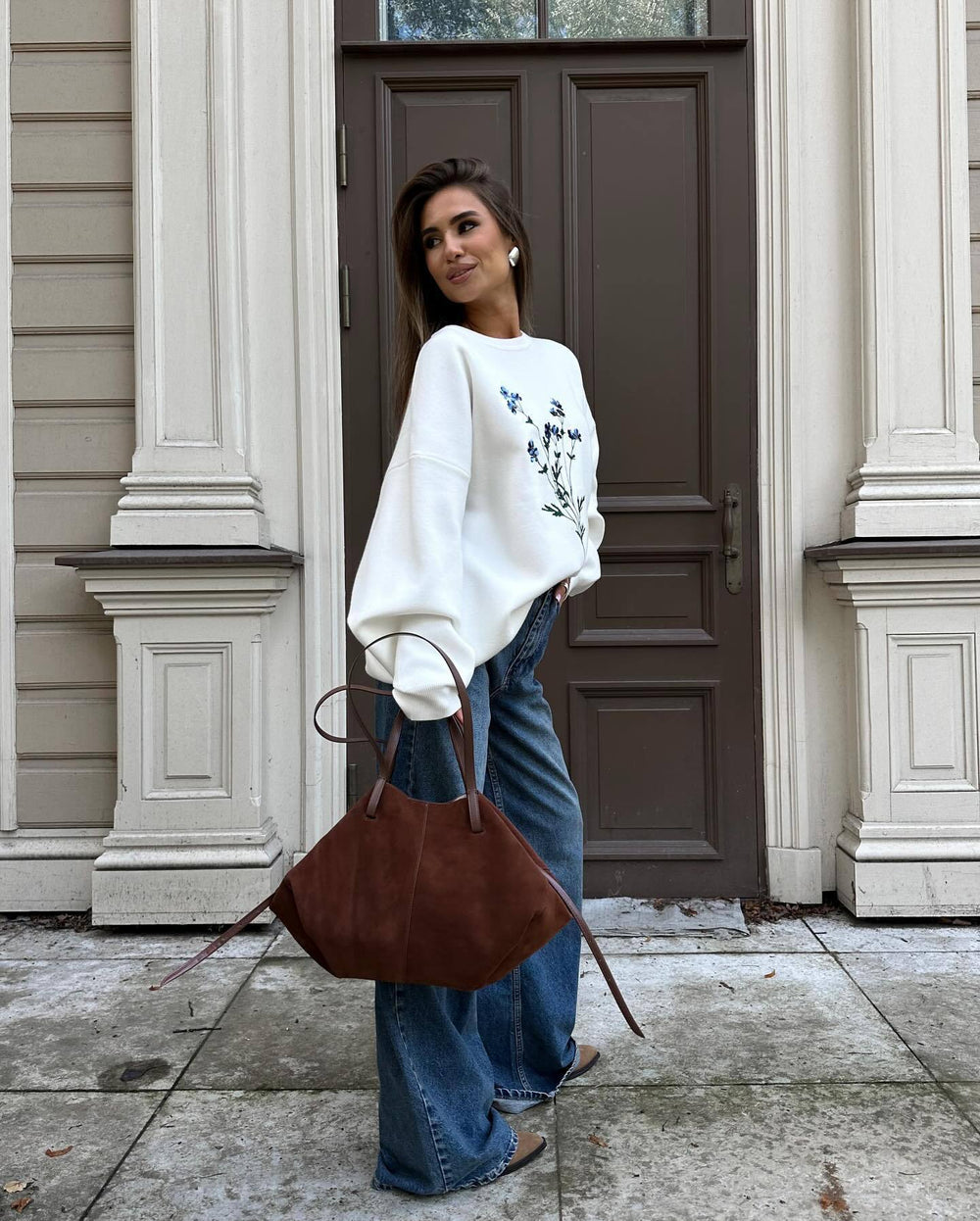 Woman in white sweater and blue jeans holding a brown leather bag in front of a door.