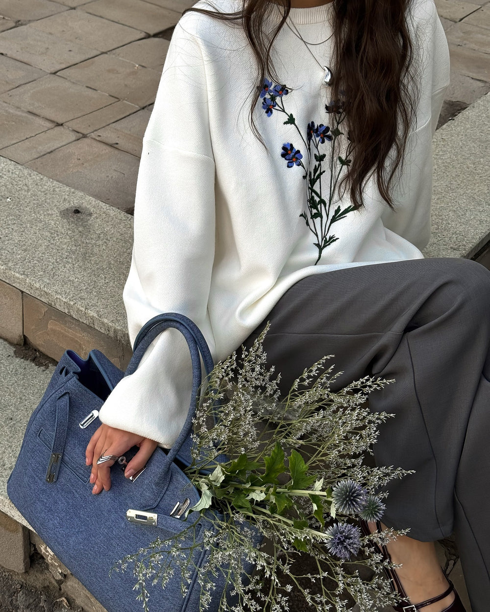 Person wearing a white sweater with floral embroidery, holding a blue handbag and a bouquet of flowers.
