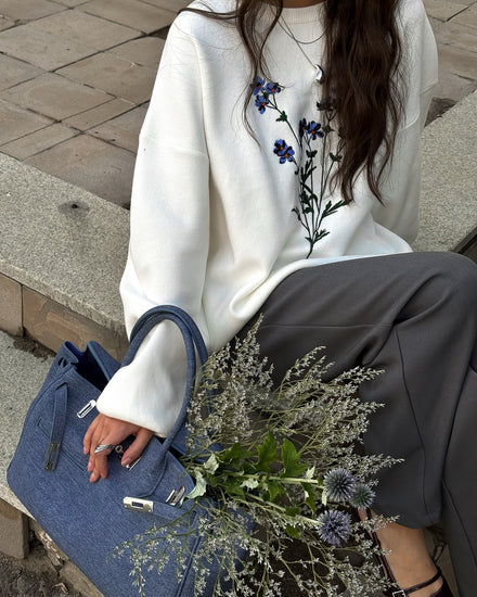 Person wearing a white sweater with floral embroidery, holding a blue handbag and a bouquet of flowers.