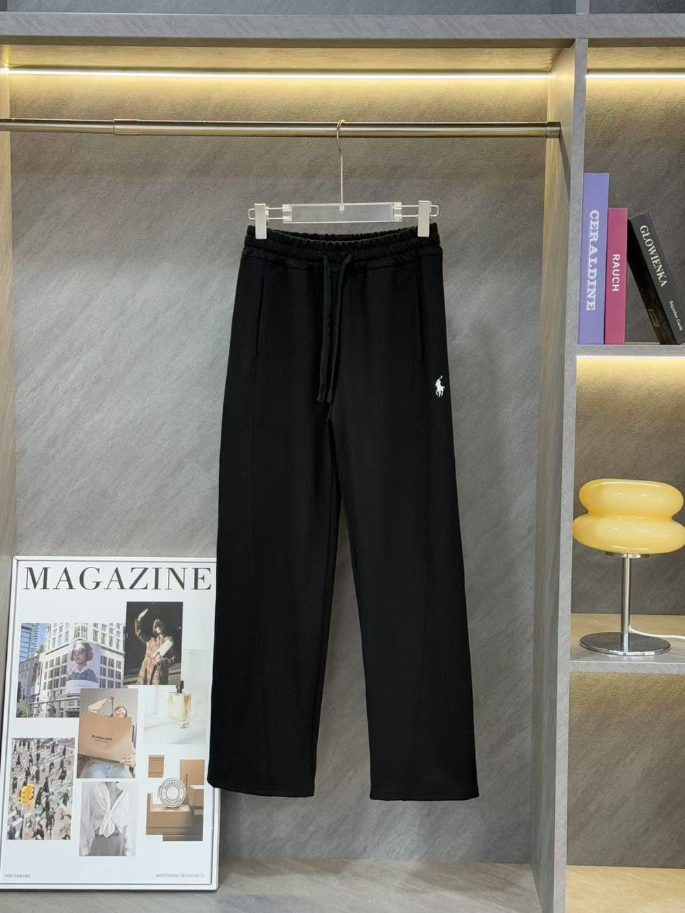 Ralph Lauren Women's Pony Straight Leg Pants. Black pants on a hanger against a gray wall with a magazine and books in the background.