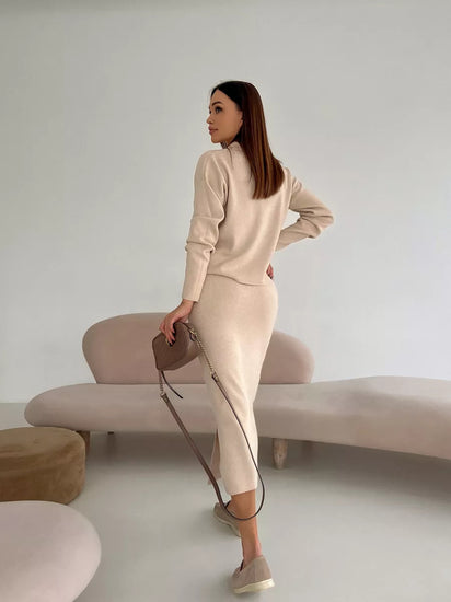 Woman in a beige outfit standing in a minimalistic room with a white wall and light-colored furniture.