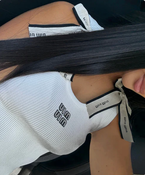 woman wearing a white sleeveless top with brand logos, sitting on a black chair.with knot