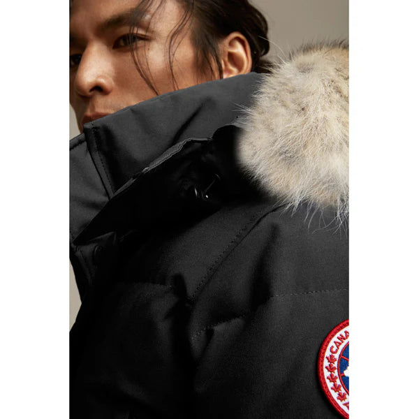 Man wearing a black coat with a fur-lined hood and Canada Goose logo. Canada Goose Wyndham Parka Goose Men's Winter Jacket at Volonka. 