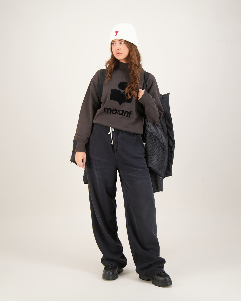 Woman wearing a dark gray sweatshirt with a logo and text, black pants, and a white beanie . Canada Goose Junction Parka Black Label Unisex Winter Jacket