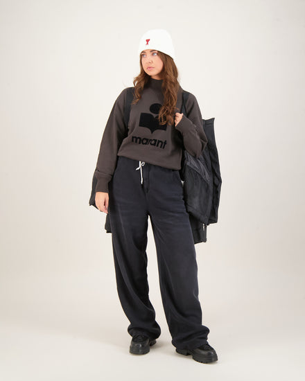 Woman wearing a dark gray sweatshirt with a logo and text, black pants, and a white beanie . Canada Goose Junction Parka Black Label Unisex Winter Jacket