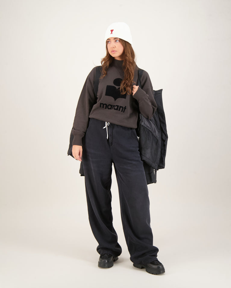 Woman wearing a dark gray sweatshirt with a logo and text, black pants, and a white beanie . Canada Goose Junction Parka Black Label Unisex Winter Jacket