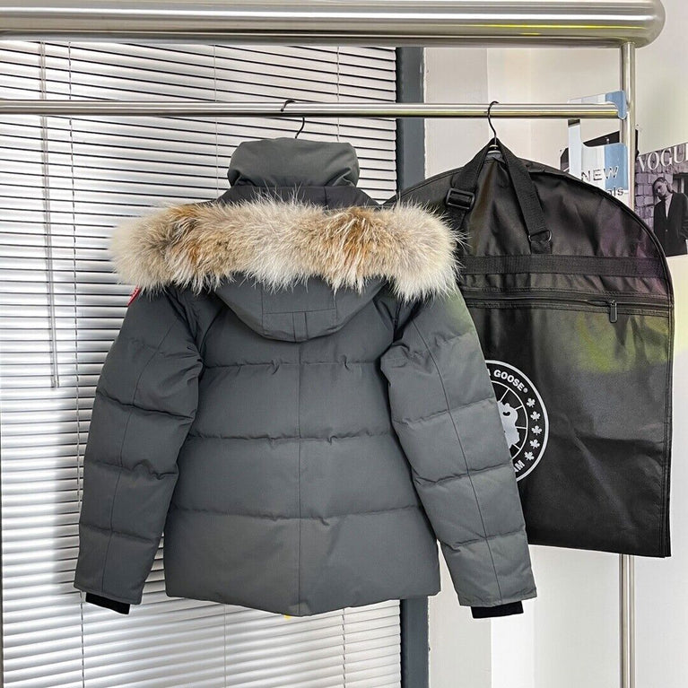 Gray puffer jacket with fur hood on a hanger next to a black garment bag with a logo. Women's/ men Canada Goose Wyndham Parka with Fur jacket
